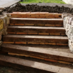 Entorno de piscina con proyecto de paisajismo rústico utilizando traviesas de tren de madera y muros de piedra natural.