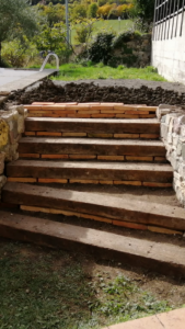 Entorno de piscina con proyecto de paisajismo rústico utilizando traviesas de tren de madera y muros de piedra natural.